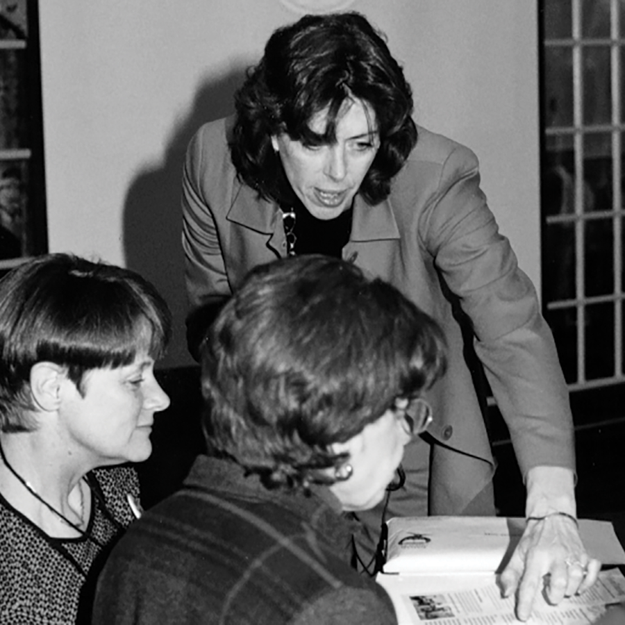 Three women are engaged in discussion at a table. One woman, standing, is pointing at a document or open book while talking to the two seated women. They are in a room with windows in the background. The image is in black and white.