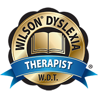 A gold badge displaying a book in the center with the words "Wilson Dyslexia" arched above and "Therapist" on a blue ribbon below. Letters "W.D.T." are at the bottom.