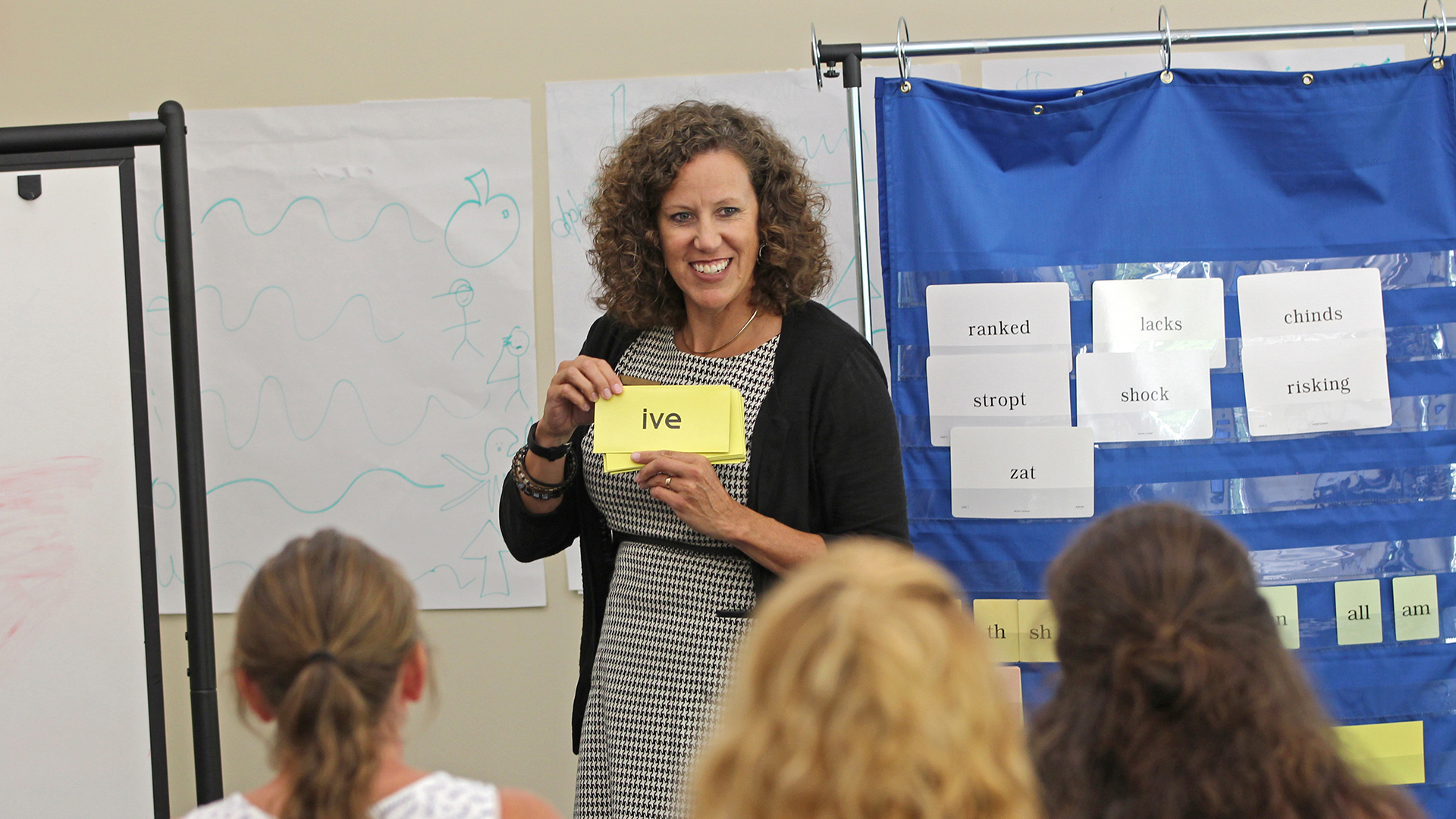 A woman with curly hair is smiling and holding a card that reads in a classroom setting. She stands near a chart with various word cards pinned on it. A whiteboard with drawings is in the background, and several people are seated and facing her.