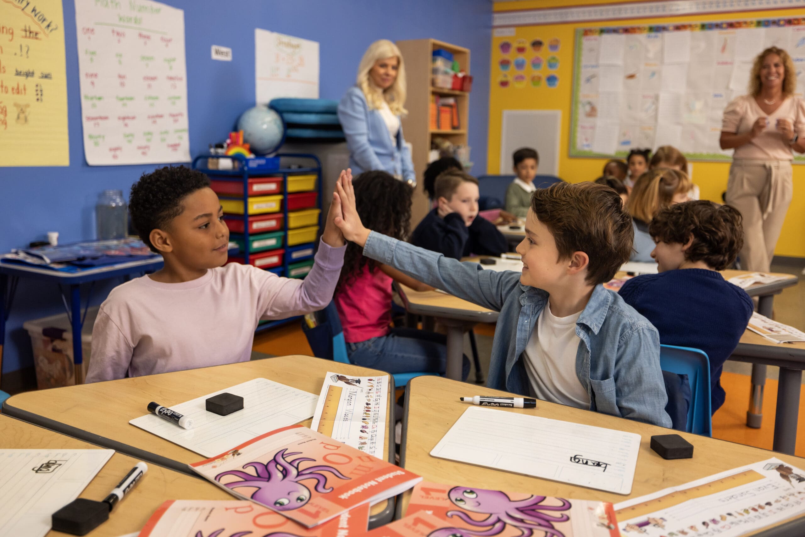Two young students high-five in a lively classroom, surrounded by desks with papers and markers. Other children and two teachers in the background engage in activities. Vibrant educational posters decorate the walls.