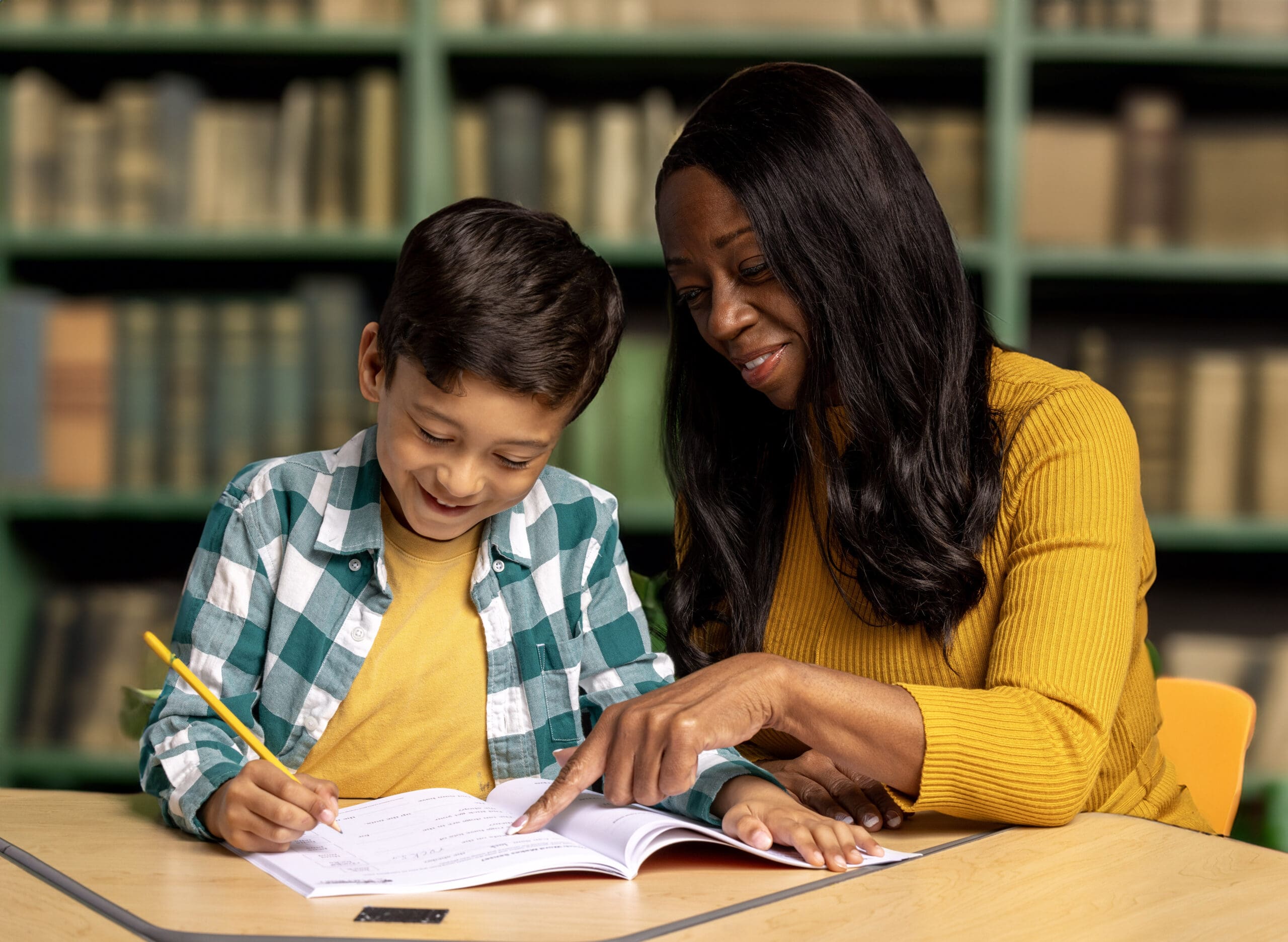 A woman and a young boy sit at a table in a library. The woman, wearing a mustard yellow sweater, is pointing at a notebook while the boy, dressed in a plaid shirt, writes with a pencil. Shelved books are blurry in the background.