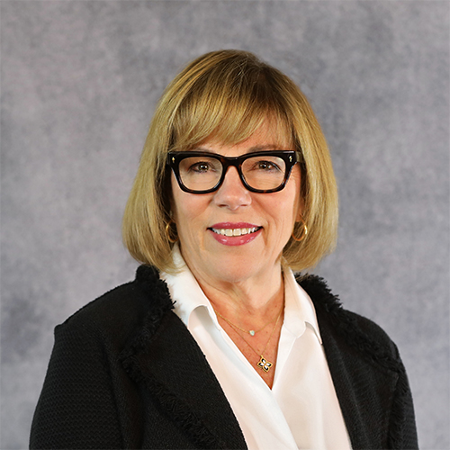 A woman with shoulder-length blond hair and black glasses smiles at the camera. She wears a white blouse under a black blazer. The background is a textured gray.