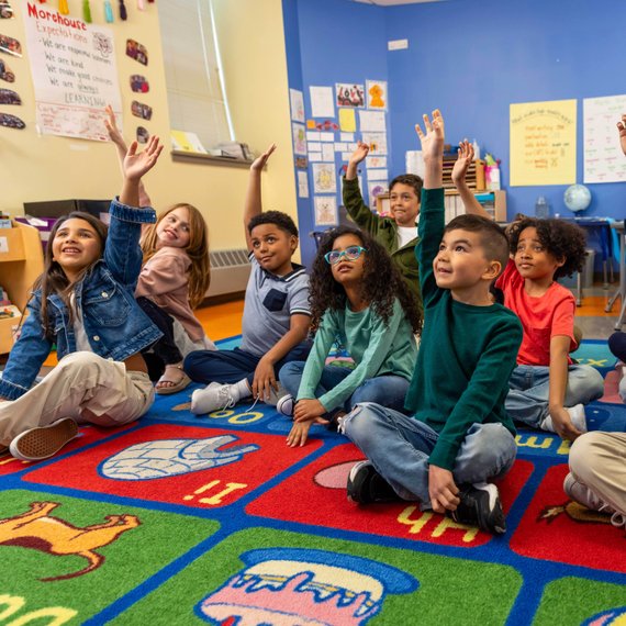 A group of young children sit on a colorful alphabet rug in a classroom, with several kids raising their hands. The walls display educational posters and a globe is visible in the background. The children appear engaged and attentive.