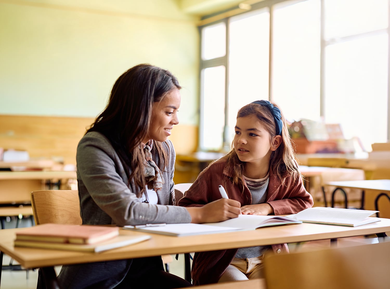 A smiling adult woman with long dark hair sits beside a young girl with a blue headband at a wooden classroom desk. The girl holds a pen and looks up at the woman while an open notebook lies in front of them. Warm natural light fills the background classroom setting.