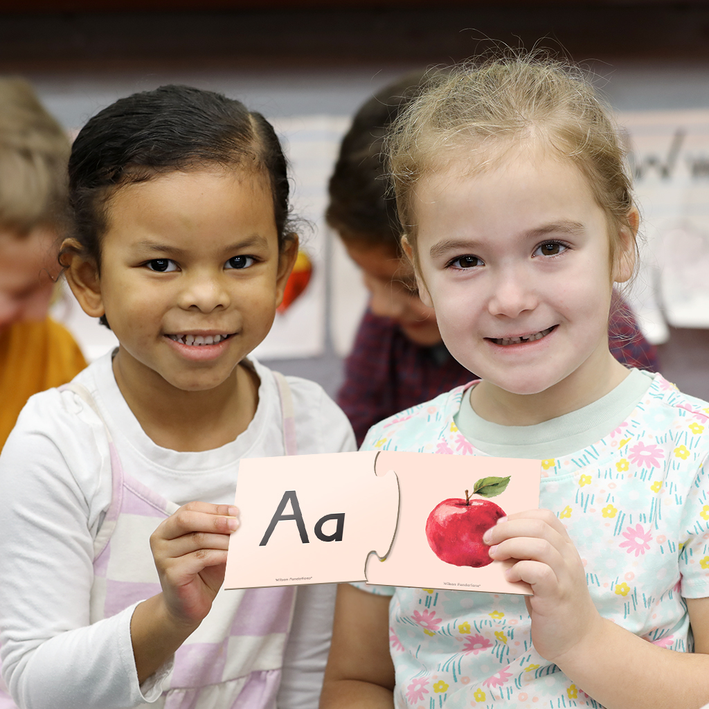 two pre-k students each holding a puzzle piece next to the other. One puzzle piece has the letter a on it while the other has an apple illustration on it.